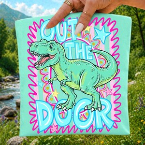 May include: A light teal tote bag featuring a cartoon Tyrannosaurus Rex in shades of green. The text "CUT THE DOOR" is displayed in a playful font, surrounded by pink and blue wavy lines and stars. The bag is held by a hand.