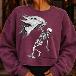 K&ouml;nnte beinhalten: Ein dunkelrotes Langarm-Sweatshirt mit einem Grafikdruck eines wei&szlig;en Skeletts, das ein flie&szlig;endes wei&szlig;es Laken mit Spinnennetz-Muster und himmlischen Details h&auml;lt. Der Text "Sheet Happens.." ist in schwarzer Schrift unter dem Skelett gedruckt.