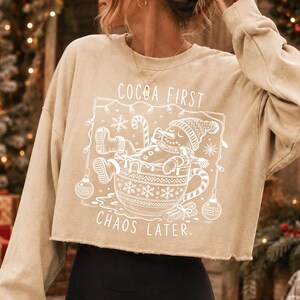 Op de afbeelding: Een beige cropped sweatshirt met een witte grafische print van een sneeuwpop die ontspant in een grote mok. De tekst luidt COCOA FIRST CHAOS LATER, omringd door een kader van kerstverlichting, sneeuwvlokken en ornamenten, met een rauwe zoom.