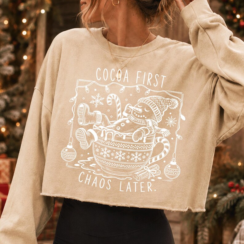 Op de afbeelding: Een beige cropped sweatshirt met een witte grafische print van een sneeuwpop die ontspant in een grote mok. De tekst luidt COCOA FIRST CHAOS LATER, omringd door een kader van kerstverlichting, sneeuwvlokken en ornamenten, met een rauwe zoom.