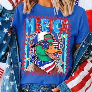 May include: Blue t-shirt with the word "MERICA" in red and blue letters. The shirt features a cartoon duck wearing star-shaped sunglasses, a baseball cap, and a bandana, all in patriotic colors. The design is framed by a red wavy border.