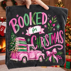 May include: A dark gray t-shirt featuring a graphic print with the text "BOOKED for CHRISTMAS" in pink and green. The design includes a pink pickup truck carrying a stack of books, a decorated Christmas tree, a pink bow, and small stars.