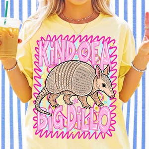 May include: A yellow t-shirt featuring a cartoon armadillo and the text "KIND OF A BIG DILLO" in pink. The armadillo is depicted in tan and pink, with a detailed shell. The text is framed by a wavy pink border.
