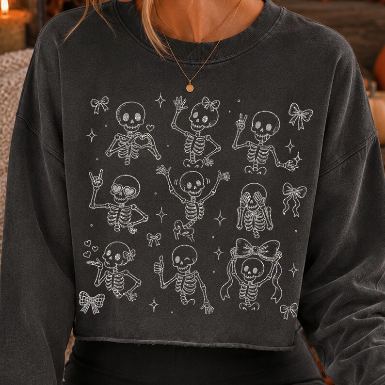 Op de afbeelding: Een antracietgrijze sweater met lange mouwen en een witte grafische print van negen verschillende skeletten in diverse houdingen. De skeletten zijn versierd met kleine strikjes, hartjes en sterretjes, wat een gedetailleerd patroon op de voorzijde vormt.