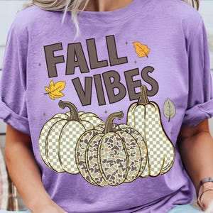 Puede incluir: Una camiseta de color morado claro con el texto FALL VIBES en letras de color marr&oacute;n oscuro. El dise&ntilde;o incluye tres calabazas con patrones de cuadros y camuflaje, acentuadas por peque&ntilde;as hojas de oto&ntilde;o amarillas y grises.