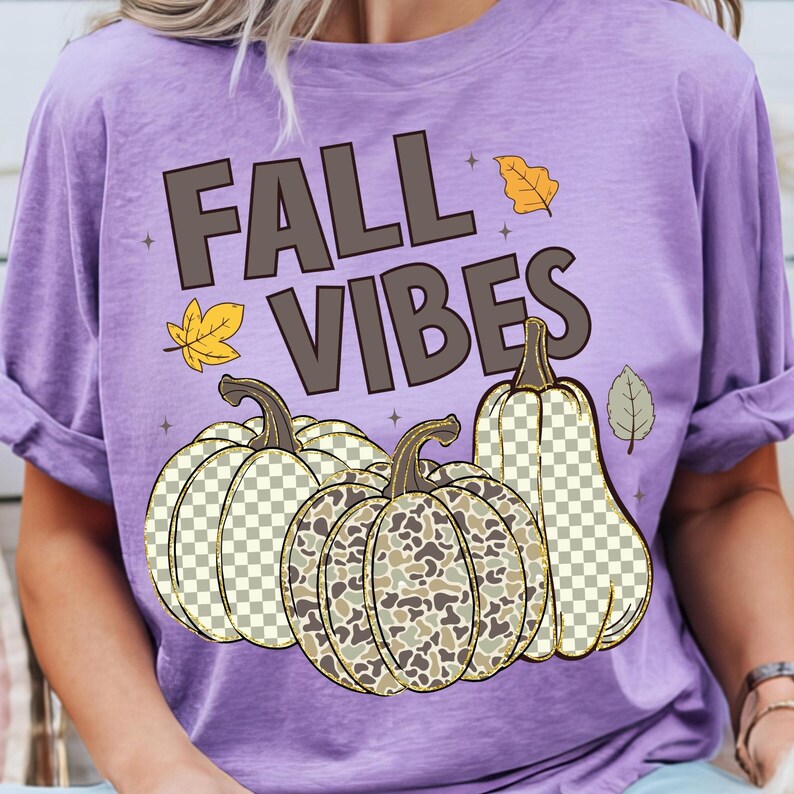 Puede incluir: Una camiseta de color morado claro con el texto FALL VIBES en letras de color marr&oacute;n oscuro. El dise&ntilde;o incluye tres calabazas con patrones de cuadros y camuflaje, acentuadas por peque&ntilde;as hojas de oto&ntilde;o amarillas y grises.