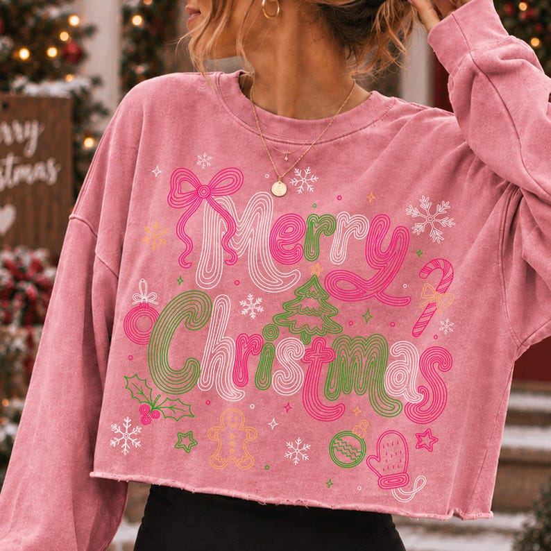 May include: A pink cropped sweatshirt featuring the text Merry Christmas in a wavy, multi-colored font. The design includes graphic illustrations of a bow, a Christmas tree, a candy cane, a gingerbread man, a mitten, snowflakes, and holly leaves.