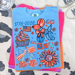 Pode incluir: Camiseta azul clara com um design patri&oacute;tico com um cupcake com uma bandeira, um animal de bal&atilde;o, cerejas e uma flor. O texto "1776-2026" e "USA" tamb&eacute;m s&atilde;o exibidos.
