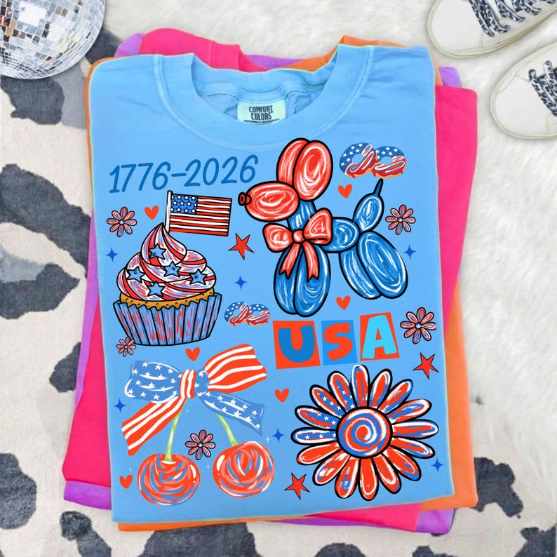 Pode incluir: Camiseta azul clara com um design patri&oacute;tico com um cupcake com uma bandeira, um animal de bal&atilde;o, cerejas e uma flor. O texto "1776-2026" e "USA" tamb&eacute;m s&atilde;o exibidos.