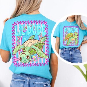 May include: A turquoise t-shirt with a cartoon turtle design and the words "Lil Dude" in a pink and blue frame. The turtle is green and brown with a smiling face. The shirt is a casual style.