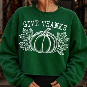 Puede incluir: Una sudadera de manga larga y cuello redondo en color verde oscuro con un estampado gr&aacute;fico blanco de una calabaza flanqueada por hojas de arce y roble. El texto "GIVE THANKS" aparece en una fuente serif blanca sobre la ilustraci&oacute;n.