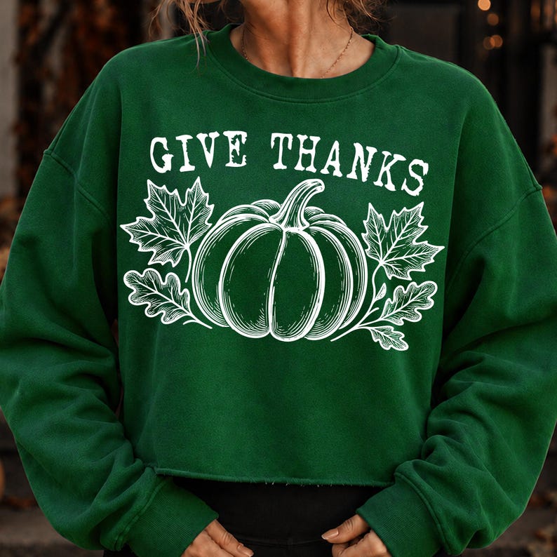 Puede incluir: Una sudadera de manga larga y cuello redondo en color verde oscuro con un estampado gr&aacute;fico blanco de una calabaza flanqueada por hojas de arce y roble. El texto "GIVE THANKS" aparece en una fuente serif blanca sobre la ilustraci&oacute;n.