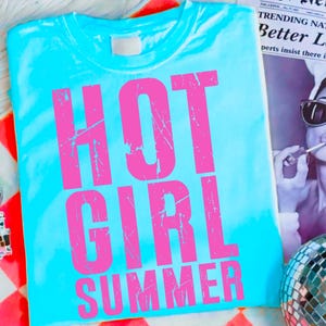 May include: A turquoise t-shirt with the words "HOT GIRL SUMMER" in distressed pink lettering. The shirt is folded and lies on a patterned surface, with a newspaper and disco ball in the background.