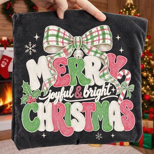 K&ouml;nnte beinhalten: Ein dunkelgraues Sweatshirt mit einem gro&szlig;en Aufdruck, der oben eine gr&uuml;n-rote karierte Schleife zeigt. Der Text Merry joyful & bright Christmas ist in stilisierter, texturierter Schrift gehalten. Das Design enth&auml;lt Stechpalmen, Beeren und eine Zuckerstange.