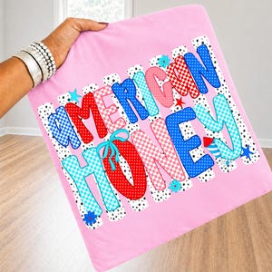 May include: A light pink square fabric item featuring the text AMERICAN HONEY in large, multi-colored, patterned letters. The letters include red, blue, and pink polka dots and checkered designs, accented with small blue and red star and flower icons.
