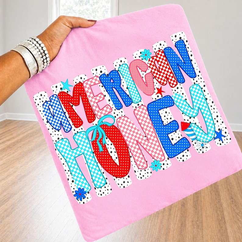 May include: A light pink square fabric item featuring the text AMERICAN HONEY in large, multi-colored, patterned letters. The letters include red, blue, and pink polka dots and checkered designs, accented with small blue and red star and flower icons.