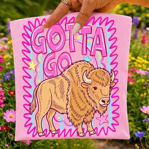 May include: Pink t-shirt featuring a cartoon bison illustration with the text "GOTTA GO" in pink bubble letters. The bison is tan with a shaggy mane and horns. The design includes stars and a wavy pink border.