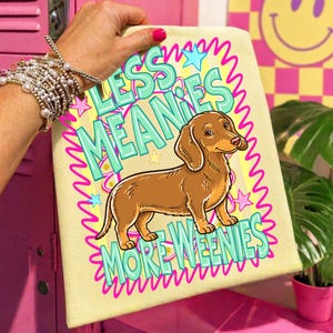 May include: A light yellow towel featuring a brown dachshund illustration. The towel has the text "LESS MEANIES MORE WEENIES" in turquoise and pink. The design is surrounded by a pink wavy border and stars.