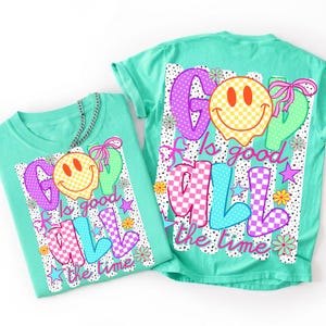 Op de afbeelding: Een mintgroen t-shirt met een grafische print met de tekst "GOD is good ALL the time". Het ontwerp bevat een geel geblokt smeltend smileygezicht, roze en blauwe geblokte letters, kleine bloemen, sterren en een roze strik.