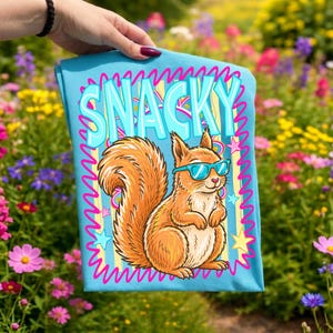 May include: A light blue t-shirt featuring a cartoon squirrel wearing sunglasses. The word "SNACKY" is printed in large, light blue letters above the squirrel. The design includes pink and yellow stars and a wavy pink border.