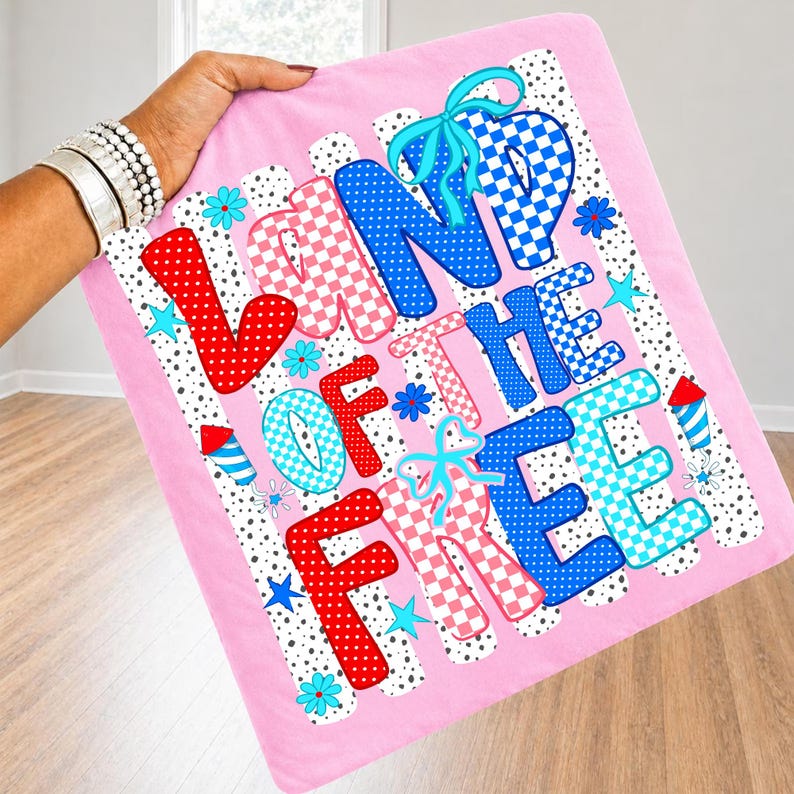 Puede incluir: Un cuadrado de tela rosa claro con el texto LAND OF THE FREE en letras grandes y multicolores con patrones de lunares y cuadros. El dise&ntilde;o incluye lazos azules, peque&ntilde;as flores turquesas, estrellas azules e iconos de fuegos artificiales.