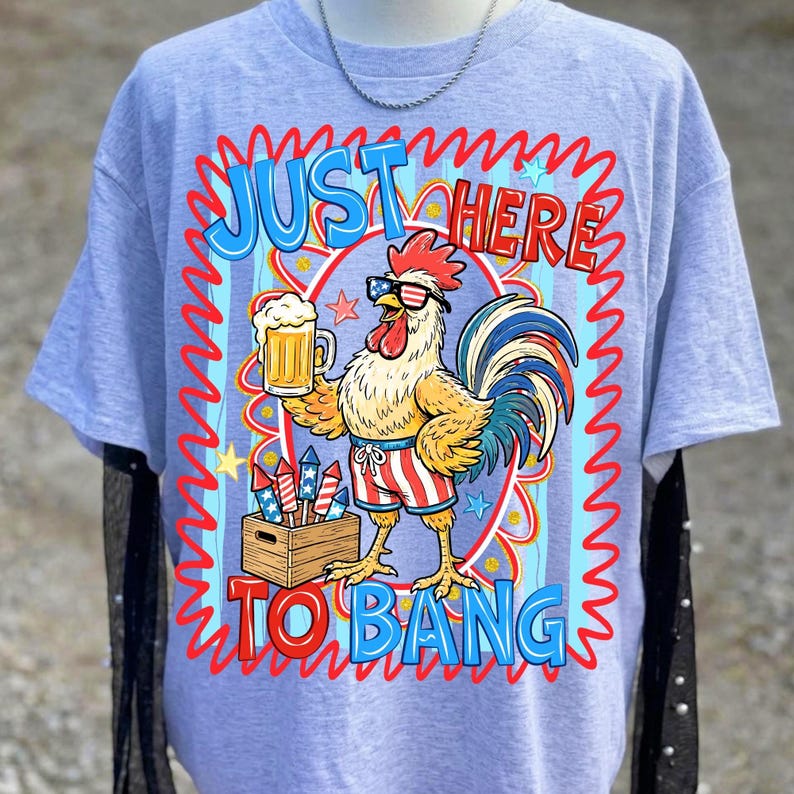 May include: Light blue t-shirt with a cartoon rooster wearing sunglasses and patriotic shorts, holding a beer mug. The shirt has the text "JUST HERE TO BANG" in a red and blue design. A wooden box of fireworks is also visible.
