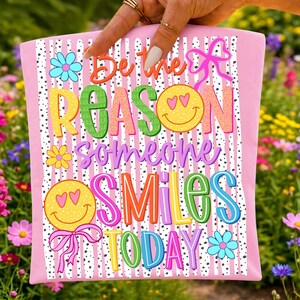 May include: Pink pillow with the text "Be the REASON someone SMILES TODAY" in colorful, playful lettering. The design includes smiley faces, flowers, and bows on a striped background.