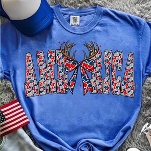 Pu&ograve; includere: Una maglietta blu con la scritta "AMERICA" in un motivo mimetico rosso, bianco e blu. Un fiocco con corna di cervo &egrave; centrato sopra la parola. Un berretto da baseball blu e bianco e una bandiera americana sono sullo sfondo.