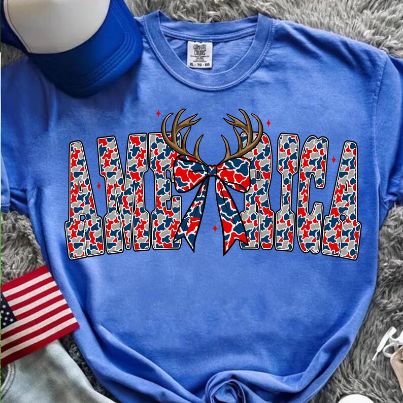 Pu&ograve; includere: Una maglietta blu con la scritta "AMERICA" in un motivo mimetico rosso, bianco e blu. Un fiocco con corna di cervo &egrave; centrato sopra la parola. Un berretto da baseball blu e bianco e una bandiera americana sono sullo sfondo.