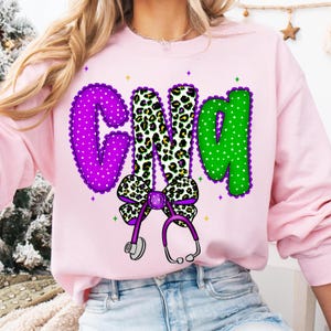 May include: Pink sweatshirt with the letters "CNA" in purple, leopard print, and green. The "C" is purple with white polka dots and a scalloped edge. The "N" is leopard print, and the "A" is green with white polka dots. A purple bow and stethoscope are below the letters.