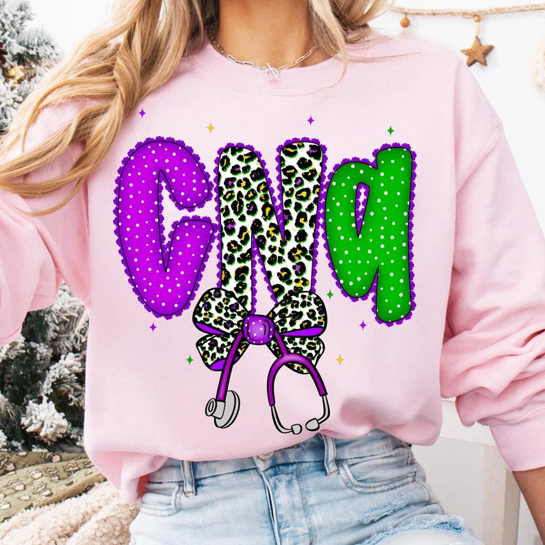 May include: Pink sweatshirt with the letters "CNA" in purple, leopard print, and green. The "C" is purple with white polka dots and a scalloped edge. The "N" is leopard print, and the "A" is green with white polka dots. A purple bow and stethoscope are below the letters.