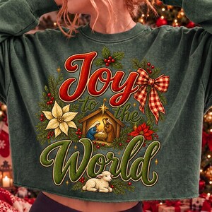 Może przedstawiać: Ciemnozielona bluza z okrągłym dekoltem z nadrukiem graficznym z napisem Joy to the World. Wz&oacute;r zawiera scenę narodzin, białą gwiazdę betlejemską, czerwone jagody, gałązki sosny, czerwono-białą kokardę w kratkę oraz małe białe jagnię.