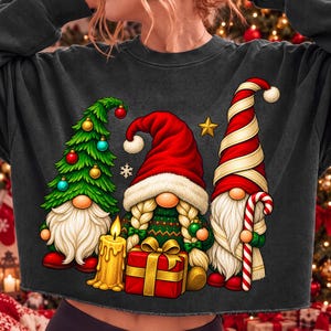 Puede incluir: Una sudadera gris oscuro de cuello redondo con un gr&aacute;fico de tres gnomos. Un gnomo tiene forma de &aacute;rbol de Navidad verde con adornos, otro tiene trenzas rubias y un gorro rojo, y el tercero lleva un gorro a rayas rojas y blancas con un bast&oacute;n de caramelo.