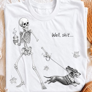 May include: A white t-shirt featuring a black and white graphic of a skeleton holding a coffee cup and walking a dachshund on a leash. The skeleton appears to be looking at the dog, with the text "Well, shit..." printed above the dog and falling leaves.