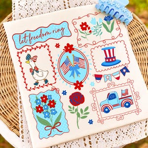May include: A cream-colored fabric featuring patriotic illustrations in red, white, and blue. Designs include a goose, flags, flowers, a truck, and the text "let freedom ring." A blue hair clip rests on the top right.
