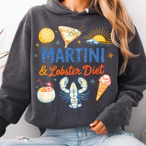 K&ouml;nnte beinhalten: Dunkelgrauer Kapuzenpullover mit einem grafischen Design mit Martini, Hummer, Eiswaffel und Kokosnussgetr&auml;nk. Der Text "MARTINI & Lobster Diet" ist in Blau und Orange gedruckt. Das Design beinhaltet eine Sonne, Sterne und eine Zitronenscheibe.