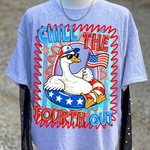 Peut inclure: T-shirt bleu clair avec un dessin d'oie de dessin anim&eacute; portant des lunettes de soleil et un chapeau patriotique, tenant un drapeau am&eacute;ricain. L'oie est dans une bou&eacute;e. Le texte "Chill the Fourth Out" est en rouge et bleu.