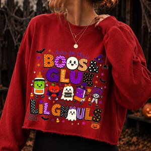 K&ouml;nnte beinhalten: Ein rotes, lang&auml;rmeliges, bauchfreies Sweatshirt mit einem farbenfrohen Grafikdruck und dem Text "here for the BOOS & GLUE with my LIL GHOULS". Das Design zeigt Zeichentrickfiguren wie Geister, eine Klebeflasche, einen Apfel und eine Katze sowie Flederm&auml;use.