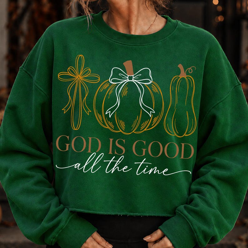 Puede incluir: Una sudadera de manga larga verde oscuro con un gr&aacute;fico de una cruz, una calabaza redonda con un lazo blanco y una calabaza alta. El texto "GOD IS GOOD" aparece en letras marrones sobre la frase en cursiva blanca "all the time".