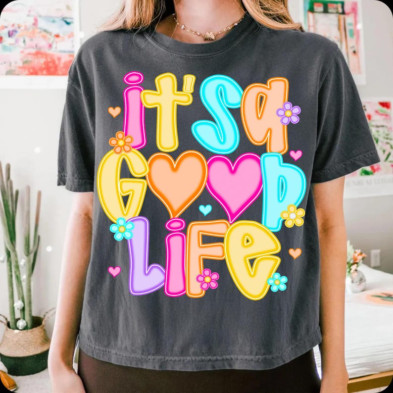 Pode incluir: T-shirt cinza escuro com a frase "It's a Good Life" em letras coloridas. O design inclui cora&ccedil;&otilde;es e flores em tons de rosa, laranja, amarelo, azul e roxo. A camisa tem um corte folgado.