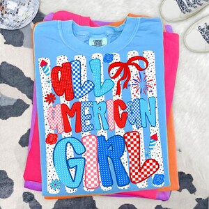 Pode incluir: Camiseta azul clara com o texto "ALL AMERICAN GIRL" em vermelho, branco e azul. O design inclui um la&ccedil;o vermelho, fogos de artif&iacute;cio e detalhes florais. A camiseta est&aacute; dobrada sobre outras camisetas coloridas.