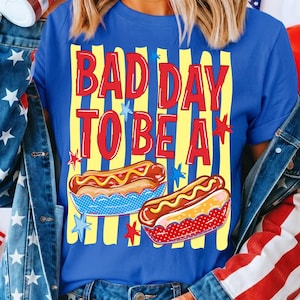 K&ouml;nnte beinhalten: Blaues T-Shirt mit dem Text "BAD DAY TO BE A" in Rot und Gelb. Das Shirt zeigt zwei Hotdogs in dekorativen Br&ouml;tchen mit Sternakzenten. Das Shirt ist ein Freizeitartikel.