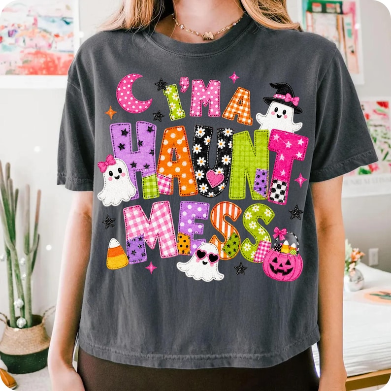 May include: A charcoal gray t-shirt featuring the colorful text "I'M A HAUNT MESS" in various patterns and colors. The design includes three white ghosts, a pink jack-o'-lantern bucket, a candy corn, and small stars on a dark fabric background.