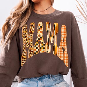 May include: A dark brown long-sleeve sweatshirt featuring the word MAMA printed in large letters across the chest. Each letter has a distinct pattern: leopard print, orange and black checkerboard, vertical brush strokes, and orange gingham.