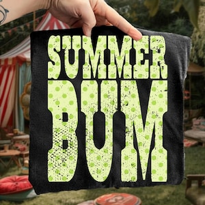 Puede incluir: Toalla negra con las palabras "SUMMER BUM" en letras grandes, desgastadas, de color verde claro con un estampado de lunares. La toalla est&aacute; sujeta por una mano, con un fondo borroso al aire libre.