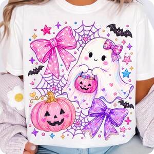 Peut inclure: Un t-shirt blanc avec une impression graphique color&eacute;e repr&eacute;sentant un fant&ocirc;me souriant tenant un seau citrouille rose, un grand n&oelig;ud rose &agrave; pois, un n&oelig;ud violet, une citrouille rose, des toiles d'araign&eacute;e, des chauves-souris noires et des &eacute;toiles.