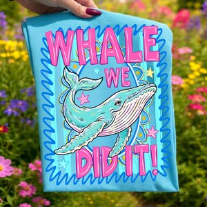 K&ouml;nnte beinhalten: Ein hellblaues T-Shirt mit einer t&uuml;rkisfarbenen Walillustration und dem pinkfarbenen Text "WHALE WE DID IT!". Das Design umfasst Sterne und einen dekorativen Rand. Das T-Shirt wird vor einem Hintergrund aus bunten Blumen pr&auml;sentiert.