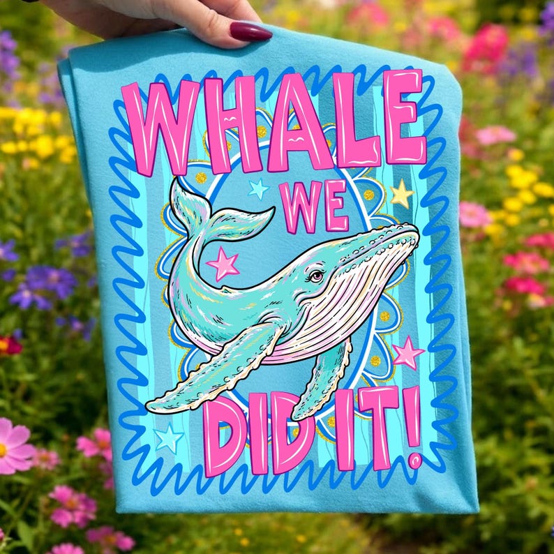 K&ouml;nnte beinhalten: Ein hellblaues T-Shirt mit einer t&uuml;rkisfarbenen Walillustration und dem pinkfarbenen Text "WHALE WE DID IT!". Das Design umfasst Sterne und einen dekorativen Rand. Das T-Shirt wird vor einem Hintergrund aus bunten Blumen pr&auml;sentiert.