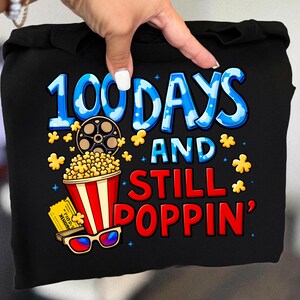 Puede incluir: Sudadera con capucha negra con un gr&aacute;fico colorido. El dise&ntilde;o presenta el texto "100 DAYS AND STILL POPPIN'" en azul y rojo, junto con palomitas de ma&iacute;z, una bobina de pel&iacute;cula, gafas 3D y una entrada de cine.