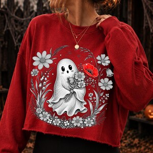 K&ouml;nnte beinhalten: Ein rotes Langarm-Sweatshirt mit einer Grafik eines wei&szlig;en Geistes, der einen Blumenstrau&szlig; h&auml;lt. Der Geist ist von einem kreisf&ouml;rmigen Arrangement aus schwarz-wei&szlig;en Strichzeichnungen von Blumen umgeben, inklusive einer roten Mohnblume.